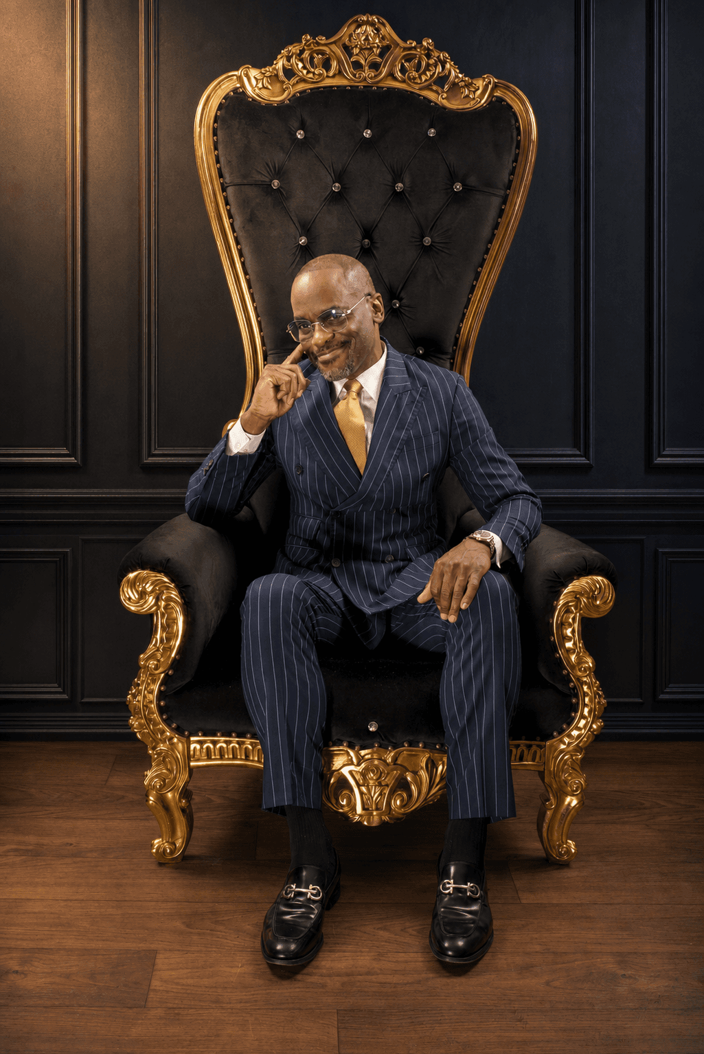 Dr. Glenn Toby in a navy pinstripe suit, seated on a black and gold throne against dark paneled walls